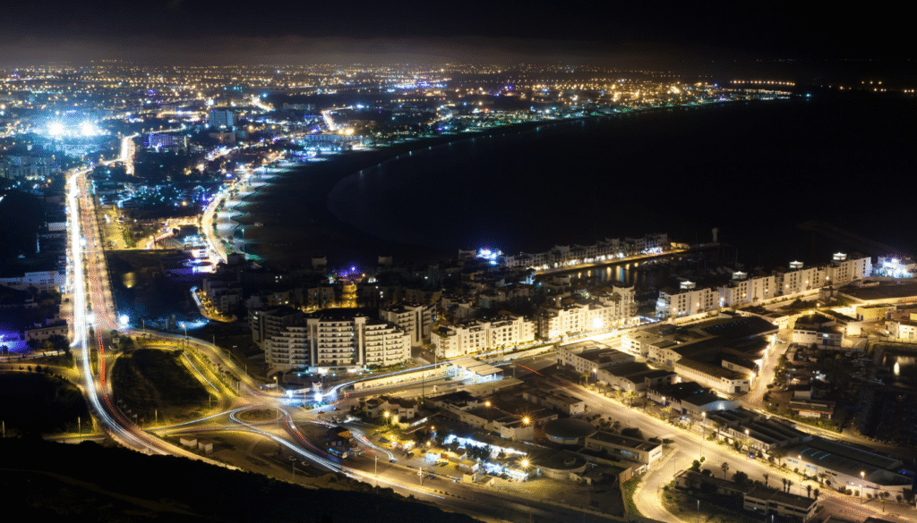 A majestic aerial view of Agadir Sports Complex, its state-of-the-art Agadir Stadium prominently featured. The multi-tiered, modern arena stands tall, its sleek, curvaceous architecture bathed in warm, golden hour lighting. The stadium's vast seating capacity is evident, with rows upon rows of comfortable seats stretching from the pitch to the upper decks. In the foreground, the lush, meticulously maintained pitch shimmers, while the surrounding concourses and facilities are visible in the middle ground. In the distance, the picturesque Moroccan landscape provides a stunning backdrop, adding to the overall grandeur of this impressive sports venue.